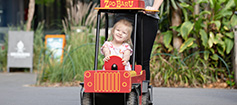 A young child in a red cart with the word 'ZooBaru' on it, being ushed by a parent.