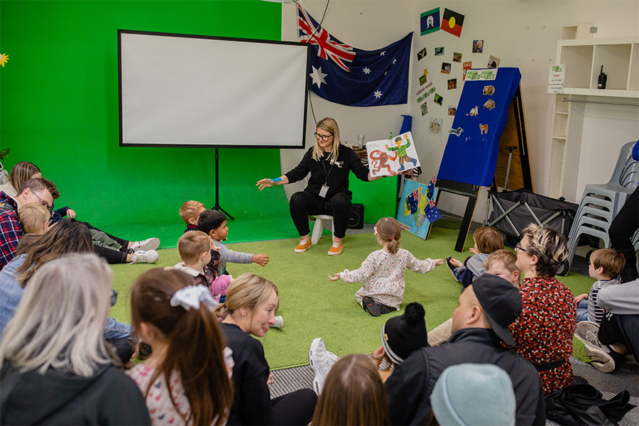 A group of children hold their arms out sitting on the ground as a teacher reads a story to them in a classroom with a green floor and walls