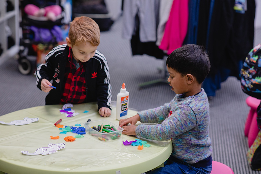 Two young boys use bottles of glue to stick coloured paper to some paper fish on a low table with a yellow tablecloth