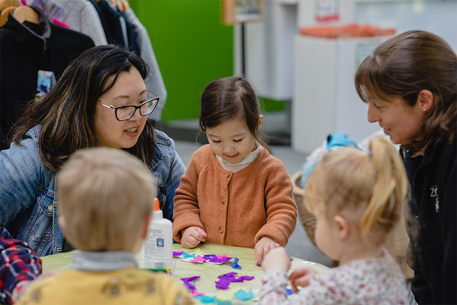 A woman watches her young daughter play with crafts on a table with other children sitting across from her