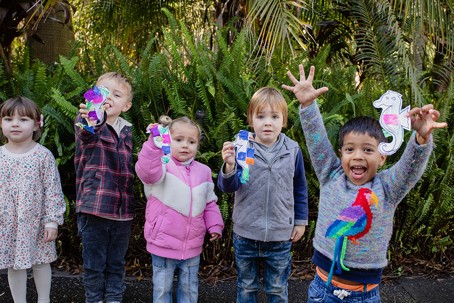 A group of young children smiling and holding up paper crafts they have made in front of leafy bushes