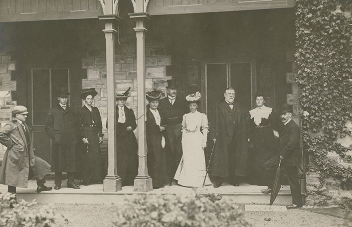 Old vintage sepia tone photograph of a group of people standing in front of an old brick building. Woman wears a white long Victorian-style dress and wide-brimmed hat. Men wear dark suits
