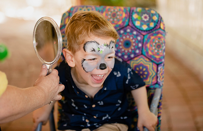 Young boy with short blonde hair laughs with koala bear face painting as woman holds up a hand mirror to his face